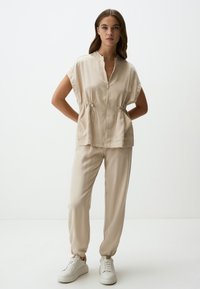 Beige Zip-Up-Oberteil mit kurzen Ärmeln und Kordelzug in der Taille, kombiniert mit passenden, schmal zulaufenden Hosen. Getragen mit weißen Sneakers. Glatte Stofftextur.