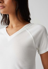 Weißes, geripptes V-Ausschnitt-T-Shirt mit kurzen Ärmeln, das einen taillierten Schnitt und eine subtile Textur aufweist. Nahtlose Schultern und weicher Stoff.