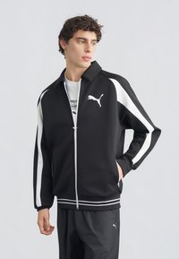 Νέος άνδρας που φοράει μαύρο και άσπρο αθλητικό μπουφάν και παντελόνι Puma, στέκεται με το ένα χέρι στην τσέπη, κοιτάζοντας στο πλάι μπροστά σε απλό φόντο.