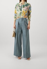 Bloemige button-up overhemd met groene, gele en roze patronen; lichte stof. Wijdvallende blauwe broek met plooien; beige handtas; lichte sandalen.