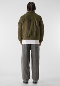 Olivgrön bomberjacka med kviltade ärmar, ribbad nederkant och avslappnad passform, matchad med stora grå jeans och svarta stövlar.