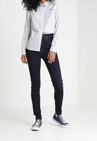 Camisa de botones ligera a rayas azules, pantalones oscuros ajustados y zapatillas navy. La camisa tiene un pequeño logo en el pecho.