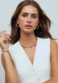 Guldmetallsmycken inkluderar: hoops örhängen, ett krökt kragnäsa och ett manschettarmband, bärs med en vit ärmlös topp.