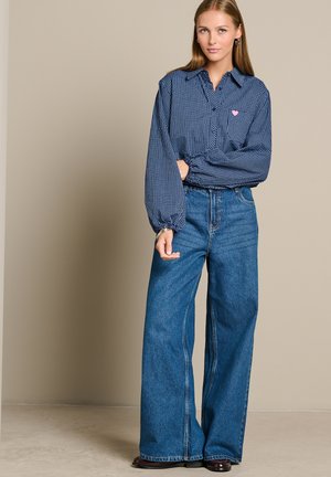 Jonge vrouw die een blauw geruit blouse met een roze hart draagt en een wijde blauwe jeans, staand tegen een beige muur.