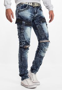 Jeans en denim bleu avec une finition déchirée, présentant des poches avec des lanières, des détails zippés et des embellissements cloutés. Accompagnés de baskets blanches.