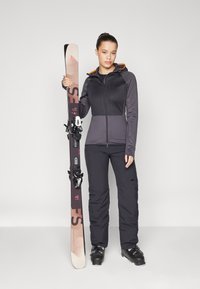 Black ski pants and a two-tone black and grey jacket with an orange hood; holding brown and black skis with bindings.