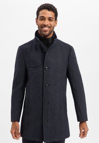 Dunkelblauer Mantel aus strukturiertem Stoff, mit hochgestelltem Kragen, vorderen Knöpfen und Seitentaschen. Weißer Hintergrund.
