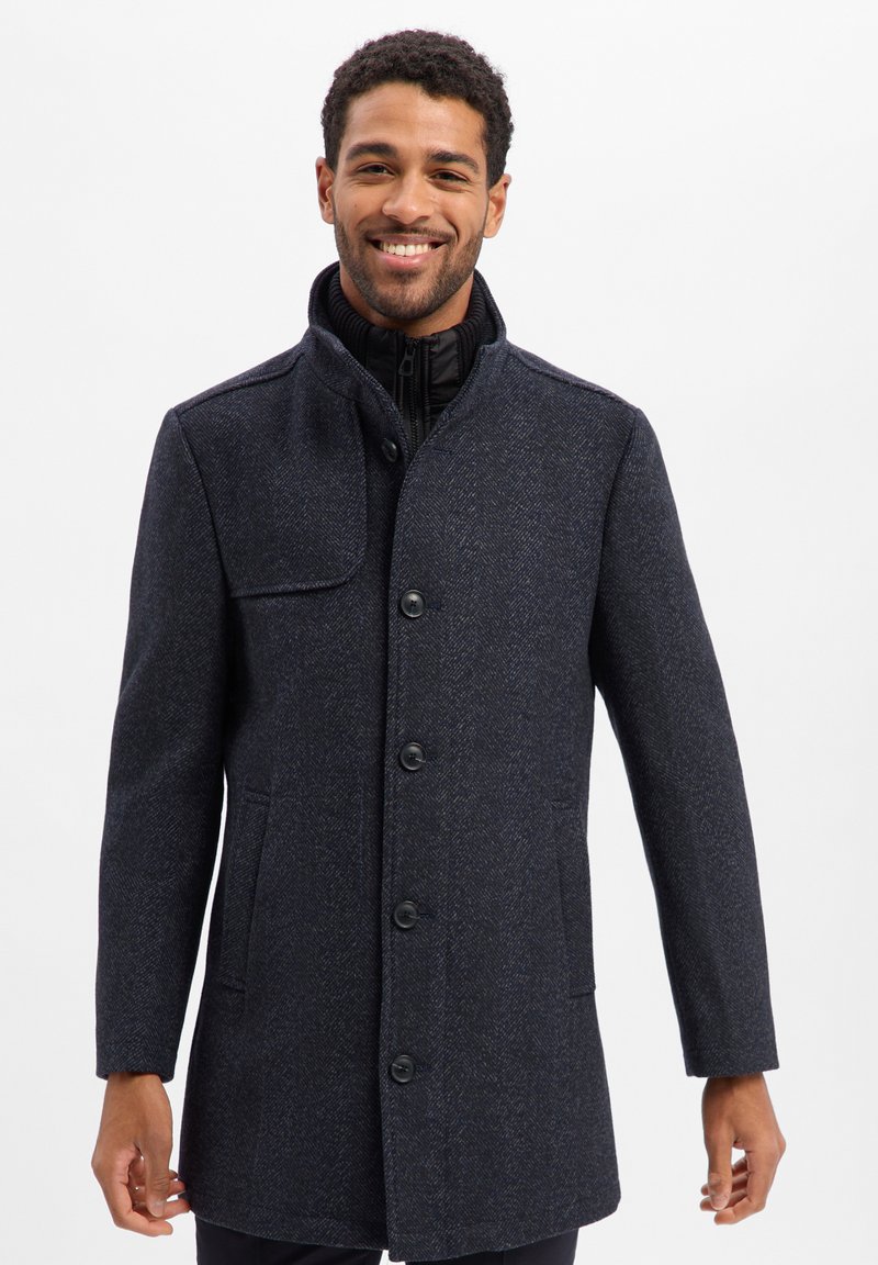 Dunkelblauer Mantel aus strukturiertem Stoff, mit hochgestelltem Kragen, vorderen Knöpfen und Seitentaschen. Weißer Hintergrund.