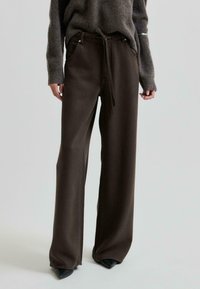 Pantaloni larghi di colore marrone scuro con vita regolabile e tasche anteriori, realizzati in tessuto testurizzato. Indossati con un maglione grigio oversize.