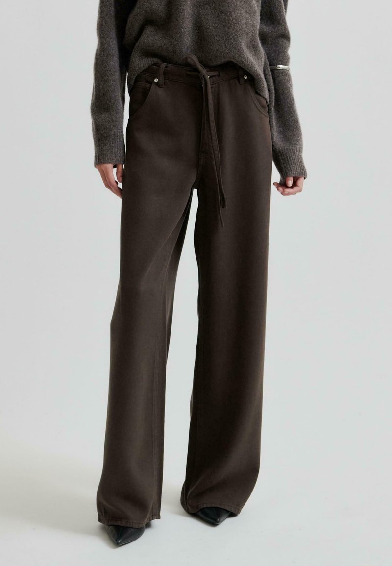 Pantaloni larghi di colore marrone scuro con vita regolabile e tasche anteriori, realizzati in tessuto testurizzato. Indossati con un maglione grigio oversize.