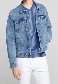 Denimjacka i ljusblått, knäppdesign med två bröstfickor och slitage. Bärs över en skjorta med blå och vita ränder.