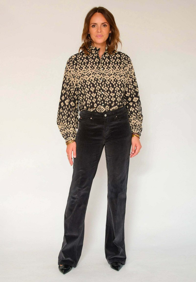 Blouse noire à motifs avec des manches longues, associée à un pantalon en velours côtelé foncé évasé. Les accessoires comprennent des boucles d'oreilles et un bracelet.