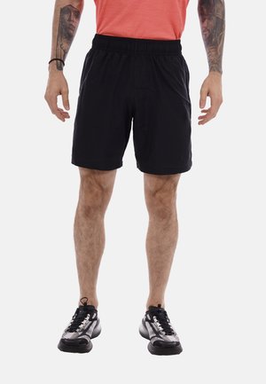 Homme portant un short de sport noir, des baskets noires et grises et un t-shirt orange, debout les bras détendus le long du corps.