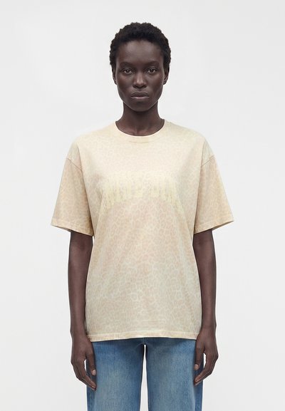 Donna con capelli corti e ricci che indossa una maglietta beige larga con stampa leopardata e jeans blu, in piedi davanti a uno sfondo chiaro e uniforme.