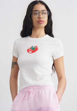 Noor naine pikkade tumedate juuste ja prillidega, kannab valget t-särki punase tomatigraafikuga ja heleroosa paelaga pükse, käed taskutes.