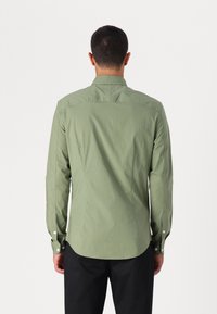 Camicia a maniche lunghe in cotone verde chiaro con yoke posteriore, orlo arrotondato e polsini abbottonati. Presenta dettagli di cucitura e una texture liscia.