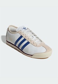 White leather sneaker with blue stripes, beige suede accents, and textured rubber sole. Features flat laces and classic branding on tongue.