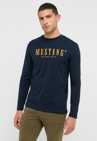 Hosszú ujjú sötétkék pamut póló, amelyen narancssárga "MUSTANG" felirat található a mellrészen, kerek nyakkivágással.