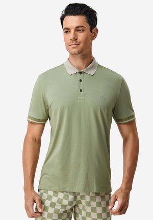 Uomo che indossa una polo verde chiaro con colletto beige e pantaloncini a quadri verdi e beige, in piedi davanti a uno sfondo bianco.