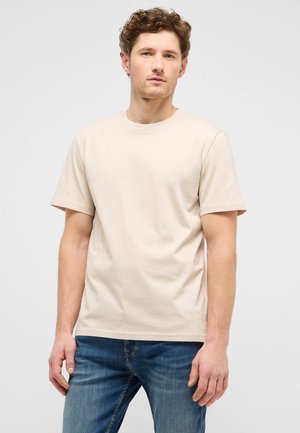 Junger Mann mit lockigem Haar, der ein schlichtes beiges T-Shirt mit kurzen Ärmeln und blaue Jeans vor einem hellgrauen Hintergrund trägt.