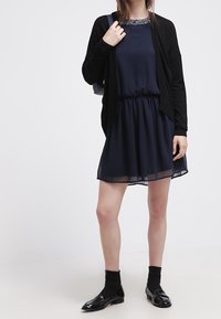 Marinsk klänning med genomskinlig kjol, samlad midja och pärlbesatt halsringning, tillsammans med en svart cardigan, svarta strumpor och blanka loafers.