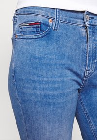 Närbild av blå jeans som bärs av en person, med framdragkedja, metallknapp, nitar och en liten ficka med ett tygdetalj.