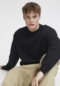 Jersey negro de punto con cuello redondo, tejido texturizado y mangas largas, combinado con pantalones beige. Modelo sentado contra un fondo blanco.