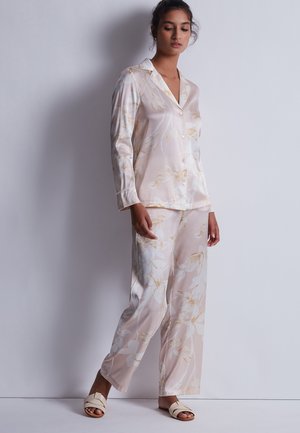 Femme debout contre un mur portant un ensemble de pyjama en satin floral rose clair et des sandales crème à bouts ouverts, regardant vers le bas avec une posture détendue.