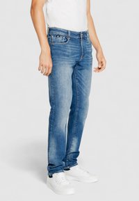 Jeans de mezclilla azul claro con un corte ajustado, que cuentan con un diseño tradicional de cinco bolsillos, un desgaste sutil y detalles de costura. Combinados con zapatillas blancas.