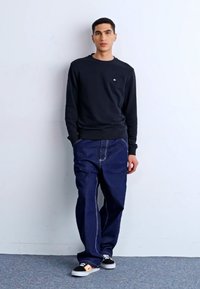 Felpa blu navy a girocollo, jeans in denim scuro larghi con cuciture bianche, scarpe da ginnastica nere con dettagli arancioni, in piedi contro un muro chiaro.