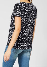 Černý top s krátkým rukávem, zdobený bílým puntíkovým vzorem, s kulatým výstřihem a volným střihem, zkombinovaný s světle modrými džíny.