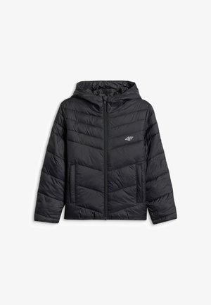 Jeune personne portant une veste matelassée noire sur un survêtement rose et une casquette de baseball rose et noire, debout avec une main dans la poche devant un fond gris.