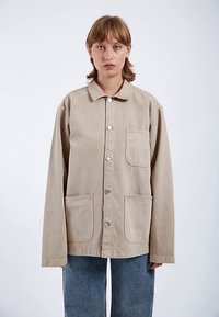 Béžová plátěná bunda s rovný střihem, velkými předními kapsami a stříbrnými patentkami. Má límec a dlouhé rukávy.