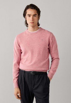 Joven con cabello oscuro que lleva un suéter rosa con cuello redondo metido en pantalones negros de talle alto con cinturón negro, manos relajadas a los lados.