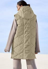 Beige Puffer-Weste mit einem Rautenmuster, übergroßer Kapuze und großem "ELBSAND"-Logo, das vertikal entlang des Rückens verläuft. Getragen über einem grauen Pullover.