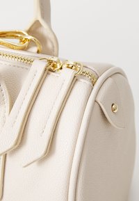 Beige textured faux leather handbag with gold hardware, featuring dual zippers and pronounced side stitching details.