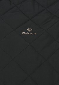 Zwarte gewatteerde stof met een ruitpatroon, met een subtiele GANT-logo in gouden borduursel nabij de bovenkant. Glad textuur en lichtgewicht ontwerp.