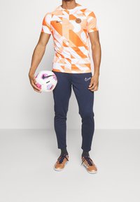 Jersey de Nike naranja y blanco con un cuello redondo, pantalones azul marino y zapatillas de fútbol marrones. La persona sostiene un balón de fútbol.
