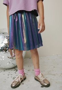 Ein schimmernder, plissierter Rock in lila, grün und blauen Farbtönen, kombiniert mit metallisch goldenen Turnschuhen und rosa gerippten Socken. In der Nähe befindet sich eine reflektierende Discokugel.