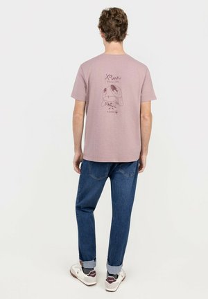 Hombre llevando una camiseta malva con un personaje de dibujos animados y texto en la parte trasera, vaqueros azules y zapatillas blancas, de pie frente a un fondo liso.