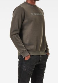 Felpa verde oliva con scollatura rotonda e polsini a coste, con logo grigio testurizzato sul petto. Abbinata a pantaloni cargo neri.