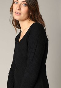 T-shirt noir à manches longues côtelé avec un décolleté en V. Le tissu présente une texture subtile et une certaine élasticité. La silhouette ajustée met en valeur la forme du corps.