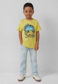 Geel katoenen t-shirt met een grafische print van bergen en groen. Lichtblauwe jeans en witte sneakers met bruinaccenten.