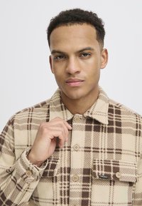 Beige and brown plaid fleece shirt with button closures and a chest pocket. Textured fabric with a collar and visible stitching details.