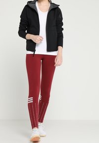 Schwarze Zip-Jacke mit grauem Futter, kombiniert mit roten Leggings mit weißen Streifen und einem strukturierten Muster. Weiße Sneakers vervollständigen den Look.
