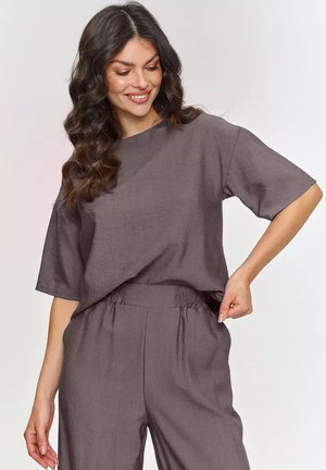 Kobieta z długimi kręconymi włosami uśmiecha się, ubrana w luźną szarą bluzkę i dopasowane spodnie z elastyczną talią, regulując pas prawą ręką.