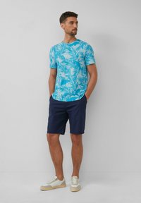 Korte mouwen teal shirt met een wit palmenbladpatroon, gecombineerd met marineblauwe shorts en lichte sneakers, staand tegen een neutrale achtergrond.