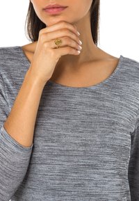 Bague en or avec un motif d'arbre, portée sur un doigt. Haut à manches longues gris marbré avec un large col. Texture lisse et coupe ajustée.