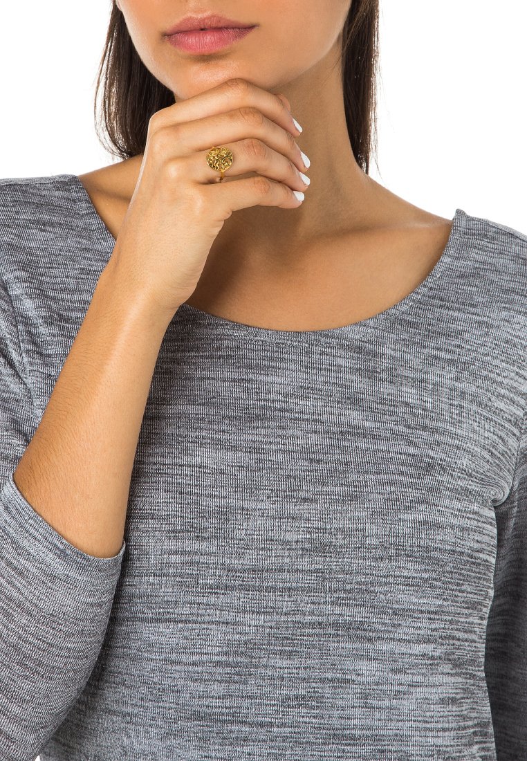 Bague en or avec un motif d'arbre, portée sur un doigt. Haut à manches longues gris marbré avec un large col. Texture lisse et coupe ajustée.