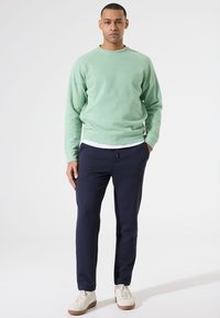 Lichtgroene sweatshirt met ribbeltextuur; marineblauwe broek; witte t-shirt eronder; crèmekleurige sneakers met lichtbruine zolen, staand tegen een witte achtergrond.